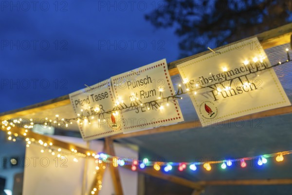 Glowing signs and fairy lights draw attention to a punch stand in the evening, Aidlingen Christmas market, Böblingen district, Germany