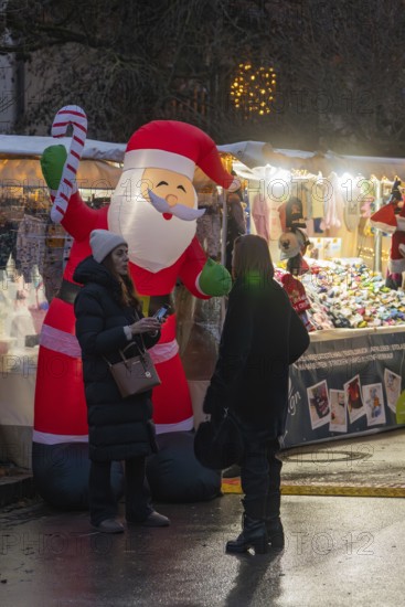 Two woman talking in front of an inflatable Santa Claus and an illuminated stand, Aidlingen Christmas market, Böblingen district, Germany