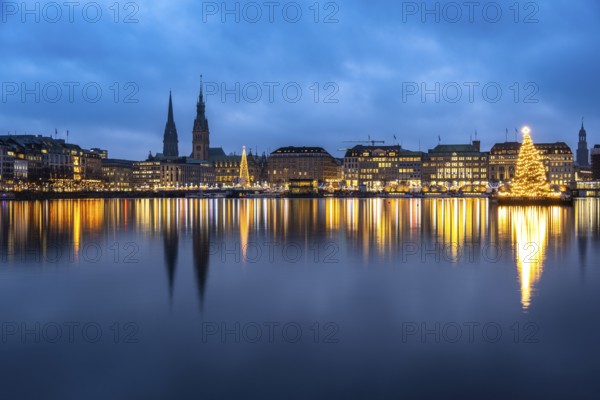 The decorated and illuminated Alster fir tree on the Inner Alster with reflections on the water in December at blue hour with the main church of St. Michaelis and town hall, Jungfernstieg, Hamburg, Germany
