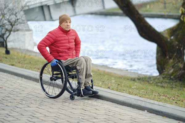 A man in a wheelchair moves along the river in the park taking a daily walk, wearing a red jacket and khaki pants