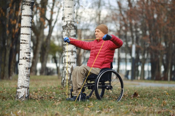 A man in a red jacket and beige hat performs strength training using resistance bands while seated in a wheelchair. He exercises outdoors in a park surrounded by trees, showcasing his determination