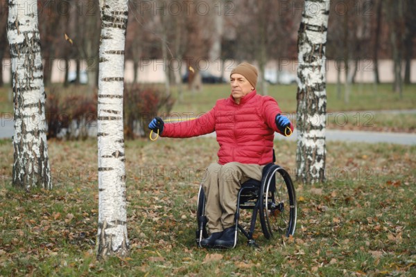 A senior man in wheelchair wearing red down jacket and gloves stretches arms outdoors in a grassy park among birch trees