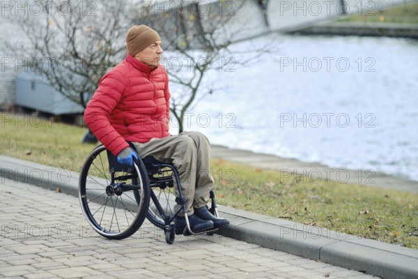 A man in a wheelchair stopped by a canal in a city park and looks at the water. He wears a red down jacket and a brown hat, enjoying the scenery as trees line the path