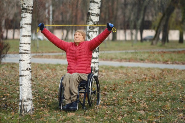 A senior old man sits in a wheelchair in a park, using a resistance band for exercise