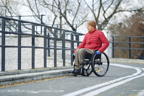 A senior man wearing a red jacket and hat is moving in his wheelchair along a smooth, winding path in a park. The trees are bare, indicating autumn, and he uses blue gloves for grip