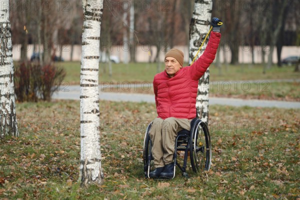 A person in a wheelchair stretches one arm up under the clear sky in a peaceful park filled with autumn leaves. The scene is serene, showcasing his determination and connection with nature