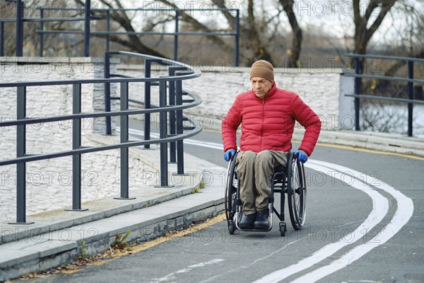An elderly man wearing a red down jacket and a hat moves steadily in his wheelchair along a curvy pathway by the river. The scene suggests a peaceful, autumn day in a public park