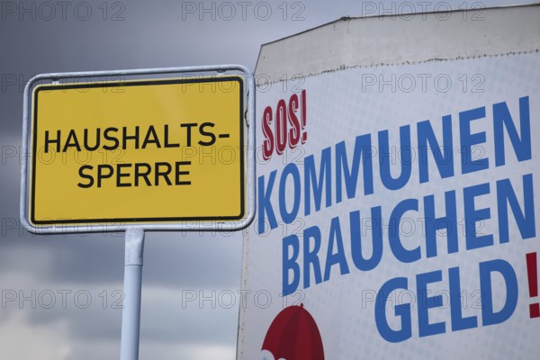 (photo montage) Town entrance sign with the inscription HOUSEHOLD LOCK next to a poster with the inscription MUNICIPALITIES NEED MONEY