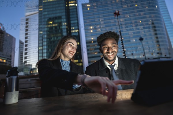 Diverse colleagues enjoying content on a digital tablet outdoors at night in an urban setting with modern skyscrapers reflecting light, sharing a moment of connection and collaborative fun