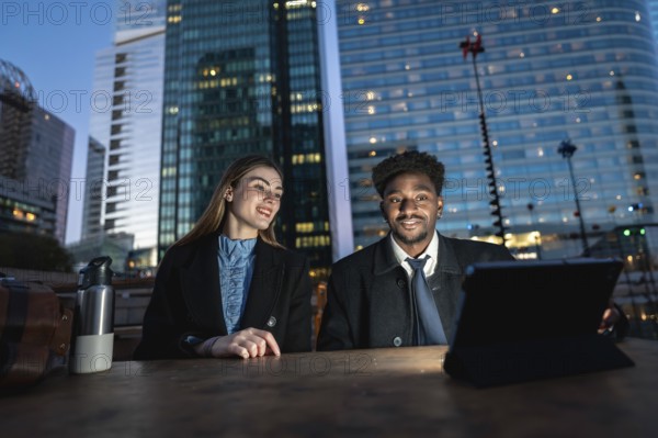 Diverse business colleagues working together on a digital tablet outdoors on a wooden table, collaborating in a city setting with illuminated modern skyscrapers in the background at dusk