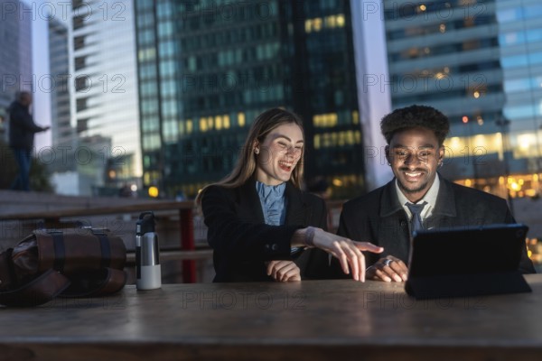 Two diverse business colleagues reviewing content on a digital tablet, discussing and laughing together after work hours in an urban setting with modern buildings