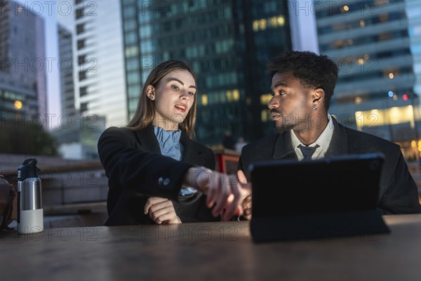 Diverse business colleagues collaborating on a project, analyzing data on a digital tablet during an outdoor evening meeting in a modern urban district, symbolizing teamwork and innovation