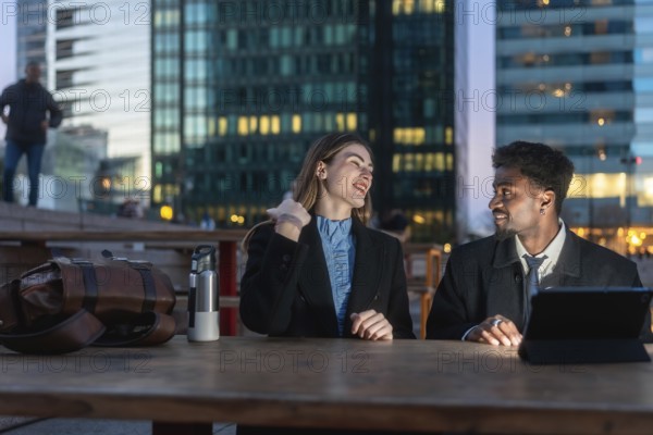 Diverse business colleagues having an outdoor conversation at dusk, sitting at a table in a modern city environment, representing teamwork, friendship, and urban professional life