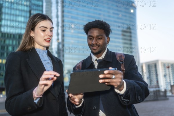 Two diverse business professionals collaborate outdoors in a modern city, smiling as they review data and strategy on a sleek tablet, sharing ideas and planning next steps