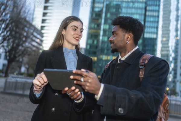Two diverse business professionals collaborating during an outdoor meeting, sharing ideas and discussing work using a digital tablet in an urban setting with modern buildings