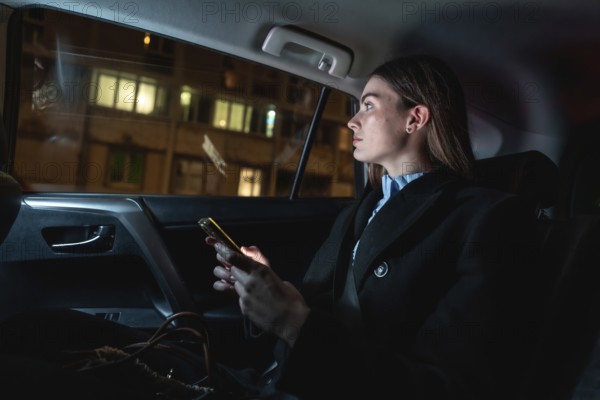 Woman traveling as a passenger in a ride hailing car at night, looking out the window while holding her illuminated smartphone, contemplating the urban cityscape passing by