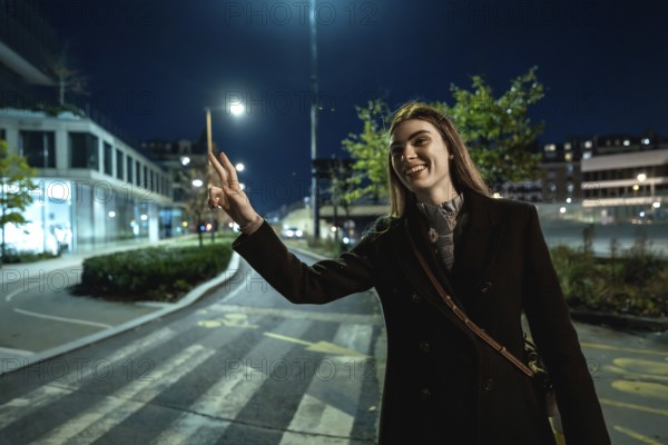 Young woman standing on an urban street at night, smiling and making a peace sign gesture while wearing a coat, conveying feelings of happiness and positivity in the illuminated city environment