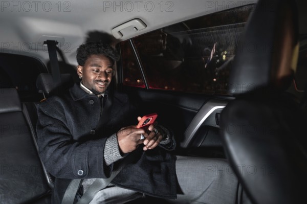 Young black man relaxed in a ride share backseat at night, smiling and browsing content on his smartphone while commuting through the city using a mobile app