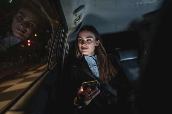 Young businesswoman commuting by car at night, sitting in backseat and looking out the window while holding a smart phone, city lights reflecting on the glass
