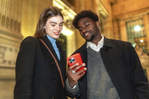 Diverse couple standing close, looking at a smartphone screen and smiling, enjoying connecting and sharing moments during a casual evening outing in the illuminated city