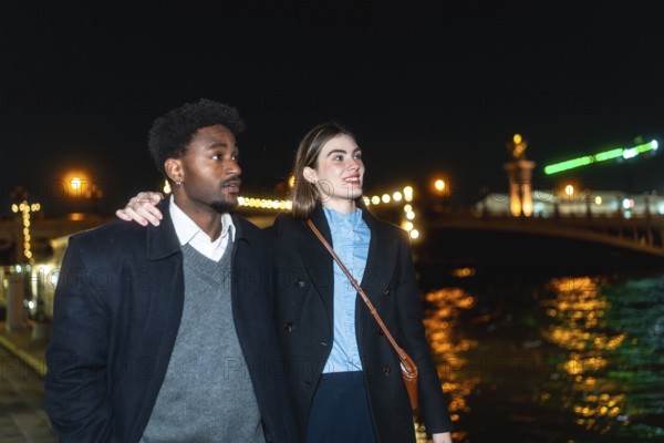 Young diverse couple strolling along a river promenade at night, city lights reflecting on the water as they hold each other, smiling and enjoying a romantic urban moment