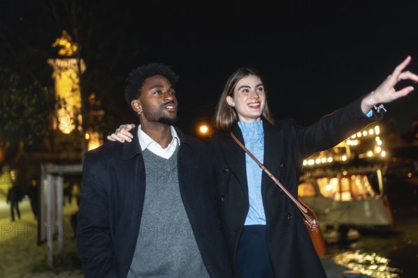 Young couple enjoying a romantic night out, with a woman pointing towards the city lights and a man looking on, sharing a moment of connection during their evening stroll
