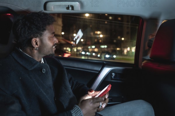 African american man in backseat of car at night smiling as he holds a glowing smartphone, looking out the window amid city lights and blurred motion during a relaxed ride share journey