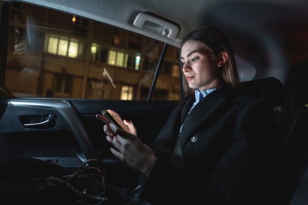 Young businesswoman commuting by car at night, focused on her smartphone as she scrolls messages and works on the go against a backdrop of city lights and reflections