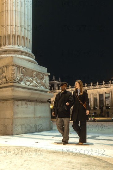 Interracial couple strolling arm in arm past an illuminated historic monument at night, sharing a romantic, candid moment of conversation and connection in the city