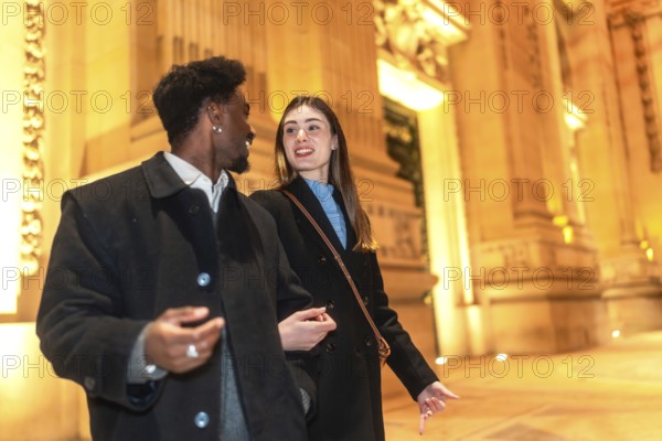 Interracial couple enjoying an evening date night, walking and talking together on a city street, illuminated by warm building lights and classic architecture