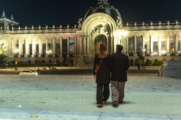 Couple walking arm in arm beneath the illuminated facades of petit palais at night, enjoying a romantic evening stroll along grand parisian streets, architecture glowing in warm light