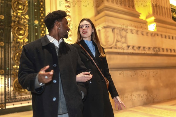 Smiling interracial couple enjoying a romantic night out, walking closely and conversing in front of a grand building with elaborate architecture, illuminated by warm lights