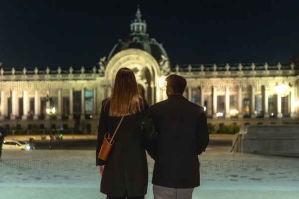 Couple is walking arm in arm, admiring an illuminated historical building with neoclassical architecture and a grand domed roof, enjoying a romantic date night in a vibrant city
