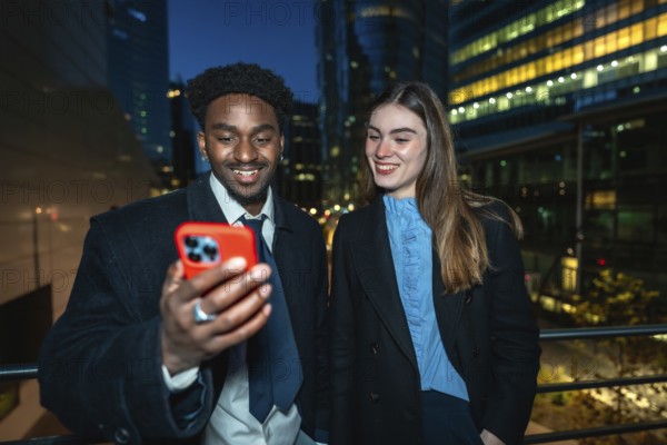 Diverse business colleagues smiling and looking at a smartphone together, enjoying a moment while connecting and sharing digital content against a modern city backdrop at night
