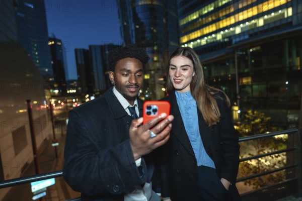 Diverse business professionals standing outside in a city at night, sharing content on a smartphone, connecting through technology and networking in an urban setting