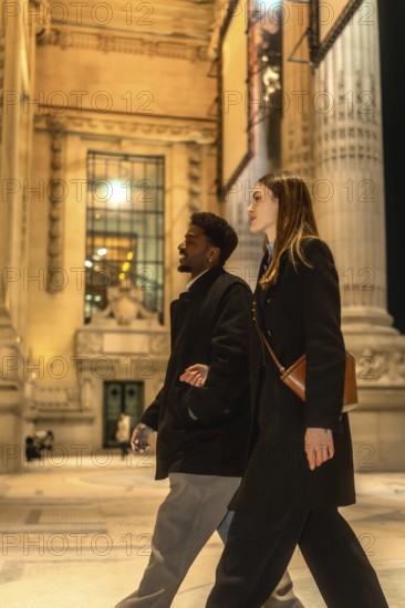 Diverse couple walking arm in arm on an illuminated city street at night, exploring urban architecture and enjoying a romantic evening stroll in downtown