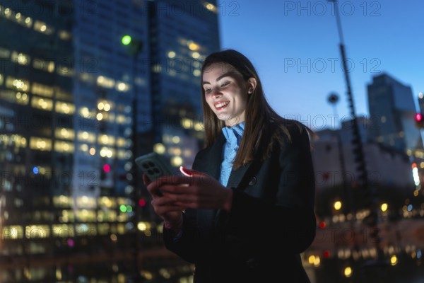 Woman smiling illuminated by smartphone screen, connecting and enjoying digital content outdoors in a modern city at night with blurred office buildings and bokeh lights in the background