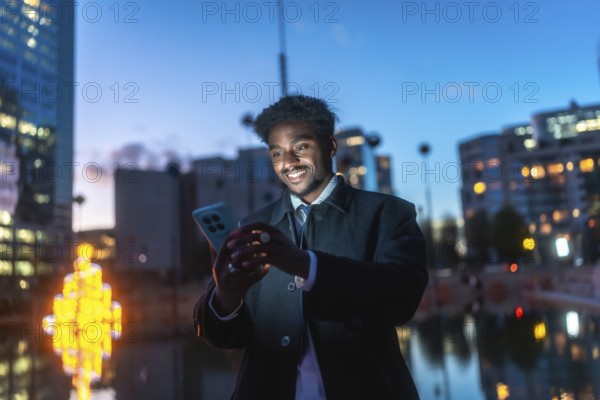 African american man smiling while engaging with a smartphone, viewing content under the illuminated city lights at night, embodying urban connectivity and modern communication