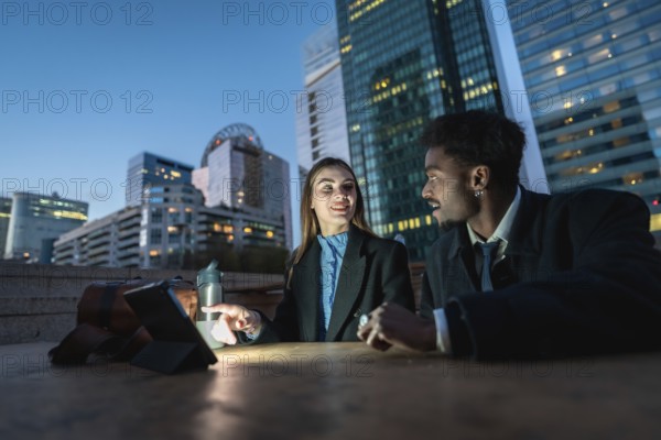 Two diverse business colleagues discussing ideas and working together on a digital tablet at night, a modern city skyline with illuminated skyscrapers framing their productive outdoor meeting