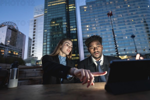 Diverse business colleagues working together on a digital tablet, pointing at the screen, with modern urban skyscrapers illuminating the evening sky in the background