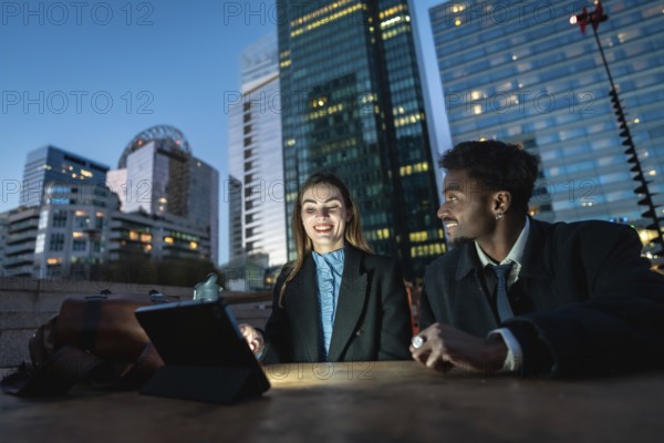 Two diverse business colleagues share a tablet at an outdoor table with a glowing downtown skyline at dusk, collaborating on digital strategy, finance and global communication