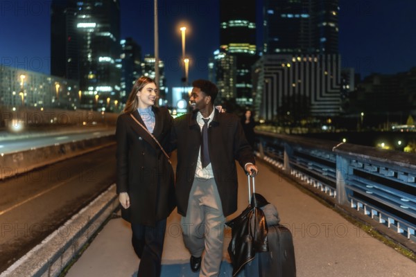Diverse couple walking together holding hands and pulling a suitcase in a modern urban city district at night, creating connected and professional travel concepts
