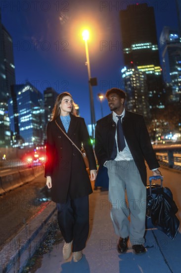 Diverse business couple walking with luggage along a downtown sidewalk at night, passing illuminated skyscrapers and a street lamp as they arrive or depart on a city trip