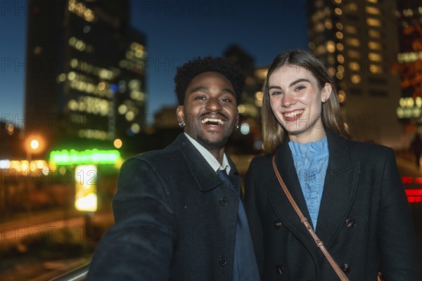 Diverse young man and woman smiling happily while taking a selfie together outdoors at night, with a modern glowing city skyline creating a soft bokeh background