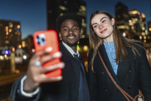 Diverse romantic couple enjoying a city date night, happily capturing their moment together with a smartphone against a blurred background of evening urban lights