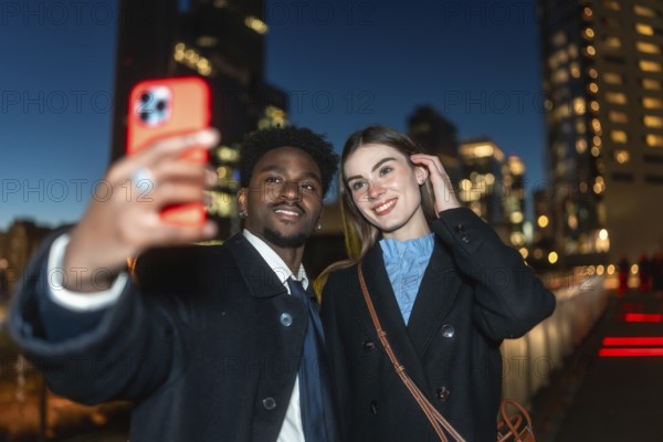 Diverse couple happily capturing a moment together using a mobile device for a selfie, illuminated by urban lights in a modern downtown area after dark