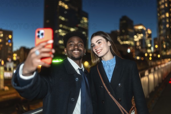 Diverse couple smiling, taking a selfie outdoors in an urban environment at night, capturing a happy moment with city lights creating bokeh in the background