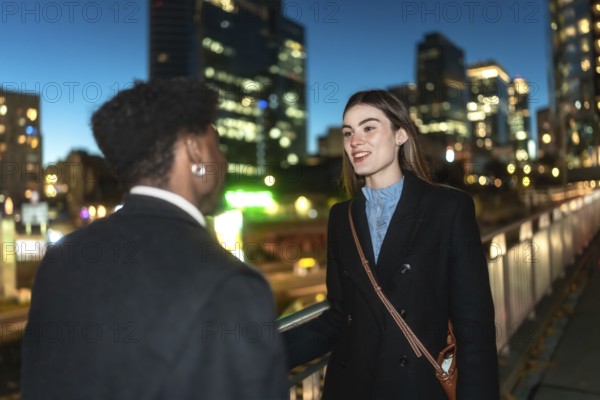 Two colleagues converse warmly on an urban pedestrian bridge at night, city lights and blurred skyscrapers in the background, suggesting networking, collaboration and modern city life