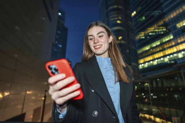 Young woman smiling outdoors at night, interacting with a smartphone in an urban environment with office buildings illuminated in the background, representing communication and social networking
