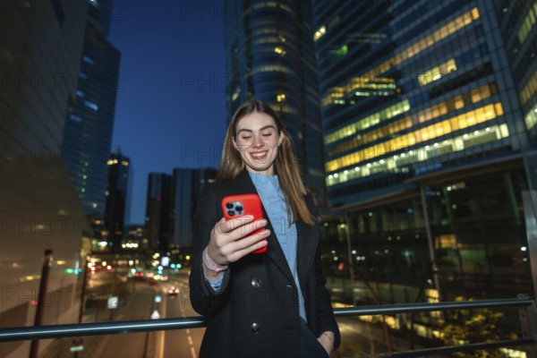 Young woman smiling while reading content on her red smartphone, enjoying digital communication and nightlife in a modern city with illuminated skyscrapers